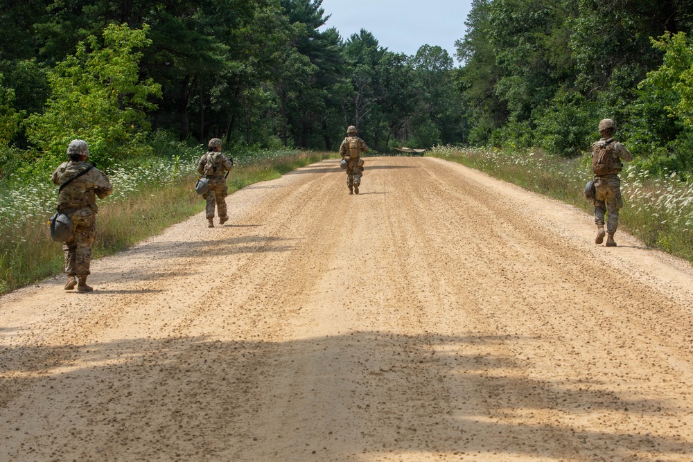 Four military personnel in camouflage uniforms are conducting a road patrol or movement exercise along a wide sandy dirt road flanked by dense green vegetation and trees. The soldiers are spread across the width of the road in a tactical formation, moving away from the camera. The setting appears to be a military training area during summer conditions.