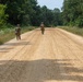 Four military personnel in camouflage uniforms are conducting a road patrol or movement exercise along a wide sandy dirt road flanked by dense green vegetation and trees. The soldiers are spread across the width of the road in a tactical formation, moving away from the camera. The setting appears to be a military training area during summer conditions.