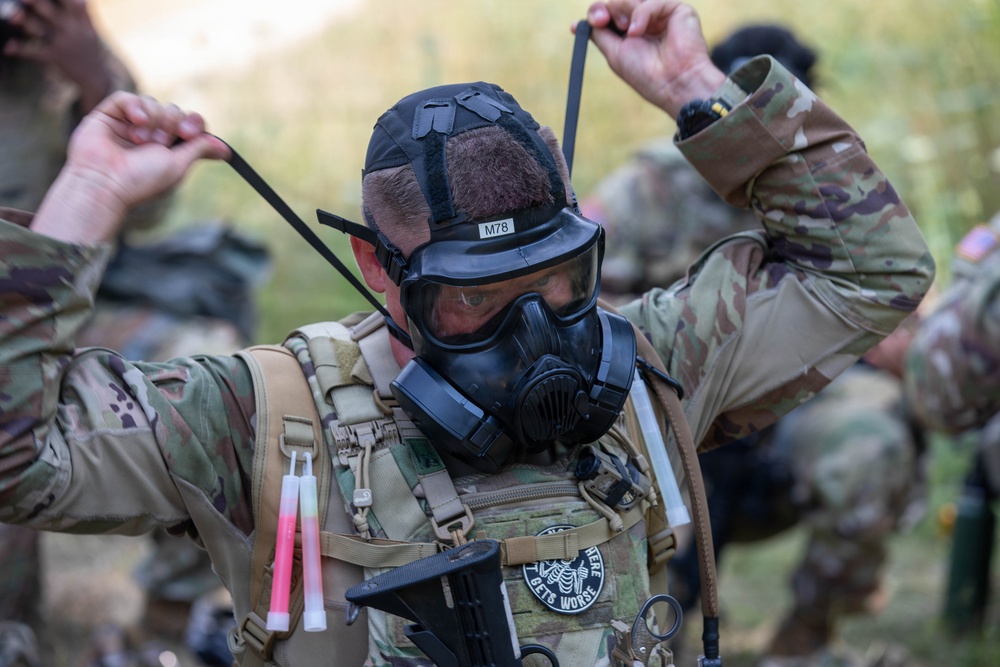 A soldier in full combat gear is donning or adjusting a military gas mask (labeled M78) in an outdoor field environment. The individual is wearing MultiCam/OCP camouflage uniform with a tactical vest, backpack, and various equipment attachments. Other soldiers in similar gear are visible in the blurred background.
