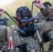 A soldier in full combat gear is donning or adjusting a military gas mask (labeled M78) in an outdoor field environment. The individual is wearing MultiCam/OCP camouflage uniform with a tactical vest, backpack, and various equipment attachments. Other soldiers in similar gear are visible in the blurred background.