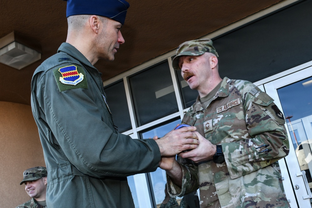 Two U.S. Air Force personnel are engaged in what appears to be a coin or award presentation ceremony outside a building. The officer on the left, wearing a flight suit with a unit patch reading 'VIDEMUS OMNIA' and a blue service cap, is presenting something to the service member on the right in OCP (Operational Camouflage Pattern) uniform with 'US AIR FORCE' tape visible. A third uniformed individual is partially visible smiling in the background.