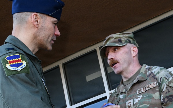 Two U.S. Air Force personnel are engaged in what appears to be a coin or award presentation ceremony outside a building. The officer on the left, wearing a flight suit with a unit patch reading 'VIDEMUS OMNIA' and a blue service cap, is presenting something to the service member on the right in OCP (Operational Camouflage Pattern) uniform with 'US AIR FORCE' tape visible. A third uniformed individual is partially visible smiling in the background.