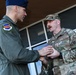 Two U.S. Air Force personnel are engaged in what appears to be a coin or award presentation ceremony outside a building. The officer on the left, wearing a flight suit with a unit patch reading 'VIDEMUS OMNIA' and a blue service cap, is presenting something to the service member on the right in OCP (Operational Camouflage Pattern) uniform with 'US AIR FORCE' tape visible. A third uniformed individual is partially visible smiling in the background.