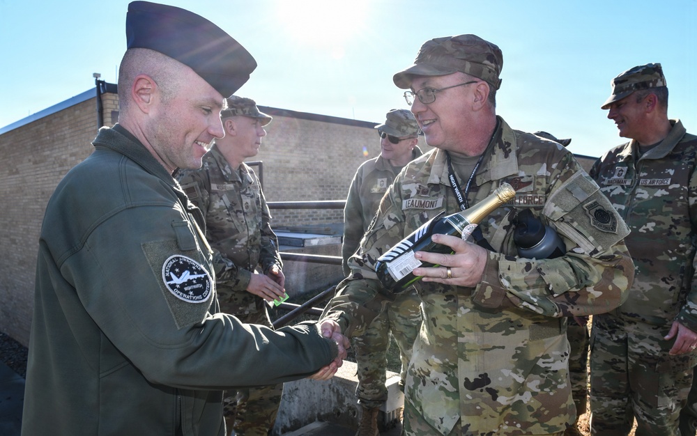 A group of military personnel in camouflage uniforms are gathered outdoors in what appears to be a ceremonial or celebratory moment. A soldier identified by a name tape reading 'LEAUMONT' is presenting a bottle of champagne while shaking hands with another servicemember wearing a dark flight suit and garrison cap. Additional uniformed personnel, including one with a 'US AIR FORCE' tape visible, are present in the background.