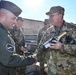 A group of military personnel in camouflage uniforms are gathered outdoors in what appears to be a ceremonial or celebratory moment. A soldier identified by a name tape reading 'LEAUMONT' is presenting a bottle of champagne while shaking hands with another servicemember wearing a dark flight suit and garrison cap. Additional uniformed personnel, including one with a 'US AIR FORCE' tape visible, are present in the background.