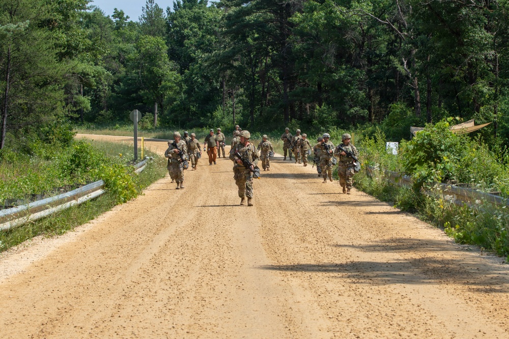 A group of military personnel in camouflage uniforms are marching along a sandy dirt road flanked by lush green vegetation and trees. The soldiers appear to be carrying weapons and gear, suggesting a training exercise or patrol movement. The scene takes place in a wooded, rural environment under bright daylight conditions.