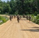A group of military personnel in camouflage uniforms are marching along a sandy dirt road flanked by lush green vegetation and trees. The soldiers appear to be carrying weapons and gear, suggesting a training exercise or patrol movement. The scene takes place in a wooded, rural environment under bright daylight conditions.