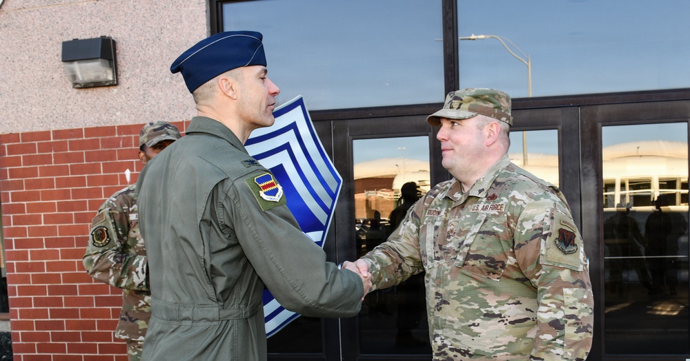 Two U.S. Air Force personnel are shaking hands in front of a building entrance, suggesting a formal greeting or recognition ceremony. The individual on the left wears an olive green flight suit with a blue garrison cap, while the individual on the right wears OCP (Operational Camouflage Pattern) uniform with 'U.S. AIR FORCE' tape visible. A large chevron insignia board is visible in the background, indicating this may be a Chief Master Sergeant-related ceremony.