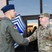 Two U.S. Air Force personnel are shaking hands in front of a building entrance, suggesting a formal greeting or recognition ceremony. The individual on the left wears an olive green flight suit with a blue garrison cap, while the individual on the right wears OCP (Operational Camouflage Pattern) uniform with 'U.S. AIR FORCE' tape visible. A large chevron insignia board is visible in the background, indicating this may be a Chief Master Sergeant-related ceremony.