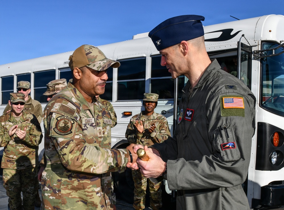 Two U.S. Air Force personnel are engaged in a coin presentation or handshake ceremony on what appears to be a flight line or base tarmac. The individual on the left, wearing OCP (Operational Camouflage Pattern) uniform with a name tape reading 'SCHULTZ,' is presenting what appears to be a challenge coin to an officer on the right wearing a flight suit and blue service cap with a colonel's eagle insignia. Several other uniformed personnel are visible in the background, some applauding.