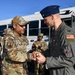 Two U.S. Air Force personnel are engaged in a coin presentation or handshake ceremony on what appears to be a flight line or base tarmac. The individual on the left, wearing OCP (Operational Camouflage Pattern) uniform with a name tape reading 'SCHULTZ,' is presenting what appears to be a challenge coin to an officer on the right wearing a flight suit and blue service cap with a colonel's eagle insignia. Several other uniformed personnel are visible in the background, some applauding.