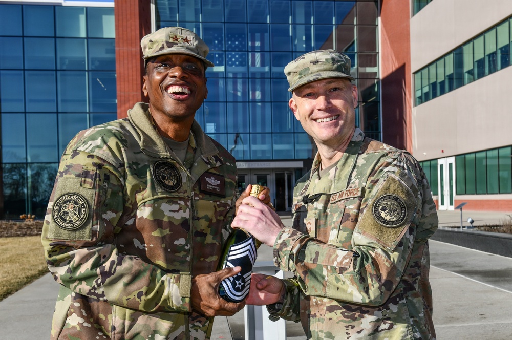 Two U.S. military personnel in OCP (Operational Camouflage Pattern) uniforms are posing together outdoors in front of a modern military building, both smiling broadly. They are jointly holding a bottle that appears to be wrapped with rank insignia, suggesting a promotion or achievement celebration. The individual on the left displays two-star rank insignia on his cap, indicating a Major General.