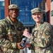 Two U.S. military personnel in OCP (Operational Camouflage Pattern) uniforms are posing together outdoors in front of a modern military building, both smiling broadly. They are jointly holding a bottle that appears to be wrapped with rank insignia, suggesting a promotion or achievement celebration. The individual on the left displays two-star rank insignia on his cap, indicating a Major General.