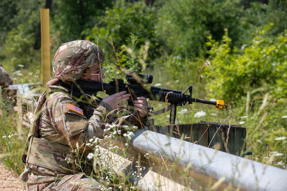 A U.S. Army soldier in MultiCam (OCP) camouflage uniform is positioned behind a wooden barrier in a field training environment, aiming an M4/M16-style rifle with an optic scope. The soldier, identifiable by the U.S. flag patch on the left sleeve, appears to be participating in a marksmanship or tactical training exercise. A yellow blank firing adapter (BFA) is visible on the muzzle of the weapon, confirming this is a training scenario.