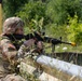 A U.S. Army soldier in MultiCam (OCP) camouflage uniform is positioned behind a wooden barrier in a field training environment, aiming an M4/M16-style rifle with an optic scope. The soldier, identifiable by the U.S. flag patch on the left sleeve, appears to be participating in a marksmanship or tactical training exercise. A yellow blank firing adapter (BFA) is visible on the muzzle of the weapon, confirming this is a training scenario.