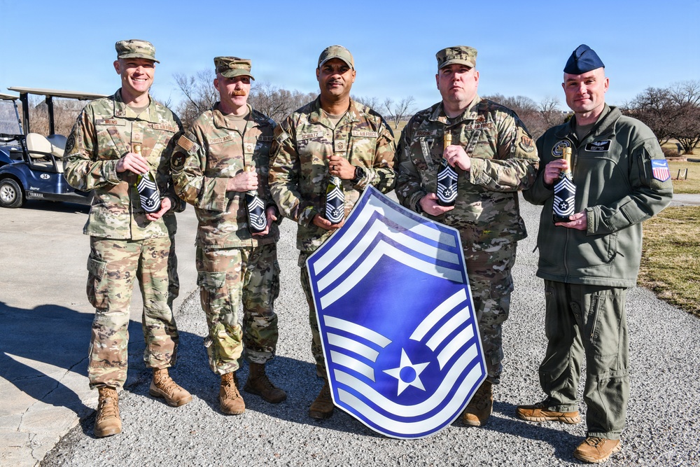 Five U.S. Air Force personnel pose outdoors holding bottles, with one individual holding a large Chief Master Sergeant rank insignia shield. The group appears to be celebrating a promotion to the rank of Chief Master Sergeant (E-9), the highest enlisted rank in the Air Force. The setting appears to be a golf course or recreational facility, suggesting an informal celebration event.