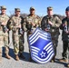 Five U.S. Air Force personnel pose outdoors holding bottles, with one individual holding a large Chief Master Sergeant rank insignia shield. The group appears to be celebrating a promotion to the rank of Chief Master Sergeant (E-9), the highest enlisted rank in the Air Force. The setting appears to be a golf course or recreational facility, suggesting an informal celebration event.