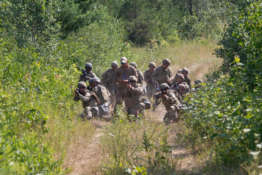 A group of soldiers in camouflage uniforms and helmets are conducting a tactical field training exercise along a dirt trail flanked by dense vegetation. Several soldiers are in kneeling or crouching positions with weapons raised, while one individual standing in the center appears to be an observer or instructor wearing sunglasses and no helmet. The scene suggests a live-fire or simulated combat training drill in a wooded outdoor environment.