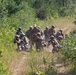 A group of soldiers in camouflage uniforms and helmets are conducting a tactical field training exercise along a dirt trail flanked by dense vegetation. Several soldiers are in kneeling or crouching positions with weapons raised, while one individual standing in the center appears to be an observer or instructor wearing sunglasses and no helmet. The scene suggests a live-fire or simulated combat training drill in a wooded outdoor environment.
