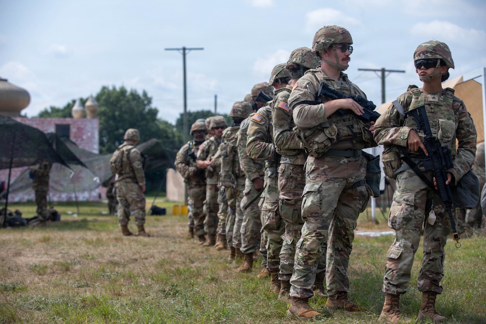 A line of U.S. Army soldiers in full combat gear stands in formation on a grassy field, likely during a training exercise. The soldiers are wearing OCP (Operational Camouflage Pattern) uniforms, helmets, and tactical vests while carrying M4-style carbine rifles. The setting appears to be a military training installation with mock structures visible in the background.