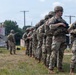 A line of U.S. Army soldiers in full combat gear stands in formation on a grassy field, likely during a training exercise. The soldiers are wearing OCP (Operational Camouflage Pattern) uniforms, helmets, and tactical vests while carrying M4-style carbine rifles. The setting appears to be a military training installation with mock structures visible in the background.