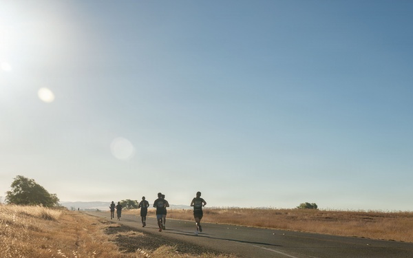 A group of runners are moving along a paved rural road surrounded by dry, golden grasslands under a bright, clear blue sky. The scene appears to be taken during early morning or late afternoon based on the low sun angle and lens flare visible in the upper left. The landscape is arid with scattered trees and rolling hills in the background.