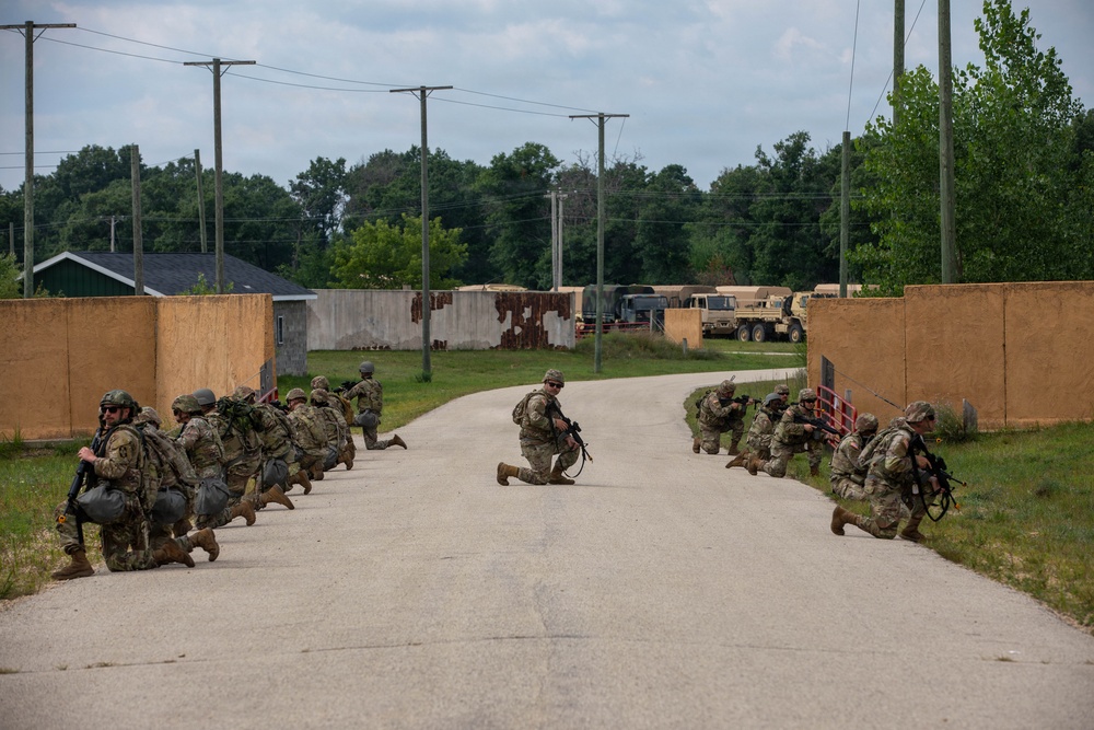 A group of military personnel in full combat gear are conducting a tactical training exercise on a paved road at what appears to be a military training facility. Soldiers are positioned in kneeling stances on both sides of the road and in the center, with weapons raised in a defensive or assault formation. The setting includes mock compound walls, utility poles, military vehicles, and surrounding tree lines consistent with a simulated urban or forward operating base environment.