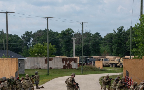 A group of military personnel in full combat gear are conducting a tactical training exercise on a paved road at what appears to be a military training facility. Soldiers are positioned in kneeling stances on both sides of the road and in the center, with weapons raised in a defensive or assault formation. The setting includes mock compound walls, utility poles, military vehicles, and surrounding tree lines consistent with a simulated urban or forward operating base environment.