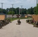 A group of military personnel in full combat gear are conducting a tactical training exercise on a paved road at what appears to be a military training facility. Soldiers are positioned in kneeling stances on both sides of the road and in the center, with weapons raised in a defensive or assault formation. The setting includes mock compound walls, utility poles, military vehicles, and surrounding tree lines consistent with a simulated urban or forward operating base environment.