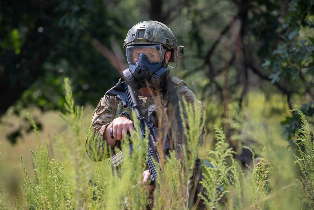 A soldier in full tactical gear is positioned in dense vegetation, wearing a gas mask and combat helmet while holding an assault rifle. The individual is partially concealed by tall green plants, suggesting a reconnaissance or patrol scenario. The setting appears to be a wooded or scrubland environment during daylight hours.