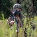 A soldier in full tactical gear is positioned in dense vegetation, wearing a gas mask and combat helmet while holding an assault rifle. The individual is partially concealed by tall green plants, suggesting a reconnaissance or patrol scenario. The setting appears to be a wooded or scrubland environment during daylight hours.