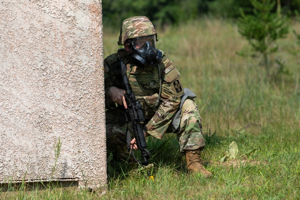 A soldier in full combat gear is crouching behind a concrete wall in a grassy, wooded area. The individual is wearing a gas mask, camouflage uniform, tactical vest, and helmet while holding a rifle. The setting appears to be a military training exercise or field operation.