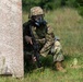 A soldier in full combat gear is crouching behind a concrete wall in a grassy, wooded area. The individual is wearing a gas mask, camouflage uniform, tactical vest, and helmet while holding a rifle. The setting appears to be a military training exercise or field operation.