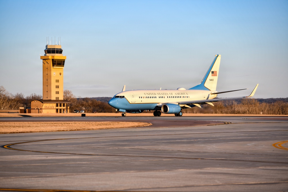 A United States of America government aircraft, appearing to be a C-40 or similar military transport, taxis on the runway at what is identified as Offutt Air Force Base (AFB). The aircraft features the distinctive white and powder blue livery associated with U.S. government/Air Force executive transport aircraft. The scene is captured during golden hour lighting with a tan control tower visible in the background.