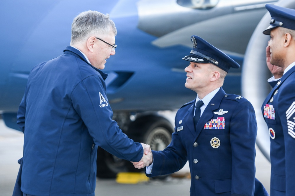 A formal military greeting is taking place on an aircraft tarmac, with a person wearing a USSF (United States Space Force) jacket shaking hands with a uniformed U.S. Air Force officer in dress blues. A third military member, appearing to be a Senior Non-Commissioned Officer, stands in the background to the right. The setting appears to be an official arrival or reception ceremony near a large military or government aircraft.