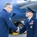 A formal military greeting is taking place on an aircraft tarmac, with a person wearing a USSF (United States Space Force) jacket shaking hands with a uniformed U.S. Air Force officer in dress blues. A third military member, appearing to be a Senior Non-Commissioned Officer, stands in the background to the right. The setting appears to be an official arrival or reception ceremony near a large military or government aircraft.