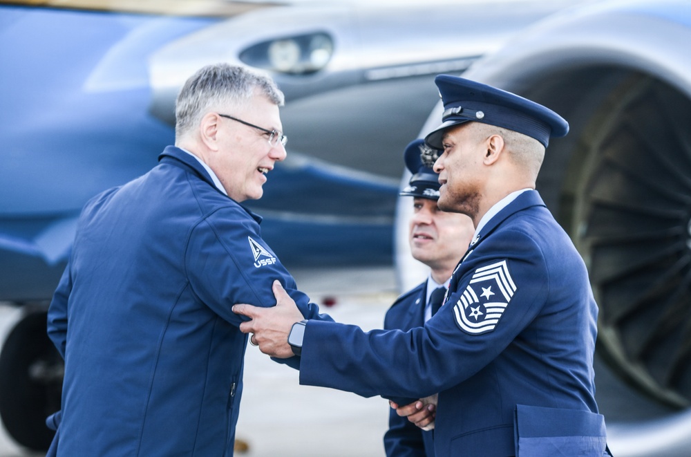 A civilian wearing a jacket with a 'USSF' (United States Space Force) patch is shaking hands with a senior enlisted military member in dress uniform near an aircraft. The military member on the right displays Chief Master Sergeant rank insignia on their sleeve. A third uniformed individual is visible in the background.