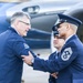A civilian wearing a jacket with a 'USSF' (United States Space Force) patch is shaking hands with a senior enlisted military member in dress uniform near an aircraft. The military member on the right displays Chief Master Sergeant rank insignia on their sleeve. A third uniformed individual is visible in the background.