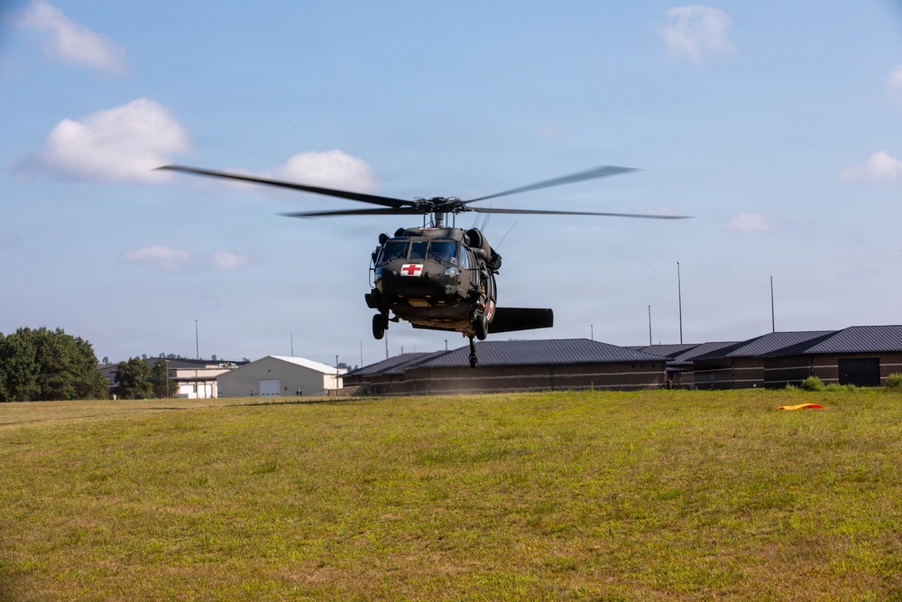 A military medical helicopter, identifiable by the red cross markings on its fuselage, is hovering or landing on a grassy field near what appears to be a military installation or airfield. The helicopter is a UH-60 Black Hawk variant configured for medevac operations. Buildings and a wind indicator (orange panel) are visible in the background.