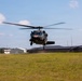 A military medical helicopter, identifiable by the red cross markings on its fuselage, is hovering or landing on a grassy field near what appears to be a military installation or airfield. The helicopter is a UH-60 Black Hawk variant configured for medevac operations. Buildings and a wind indicator (orange panel) are visible in the background.
