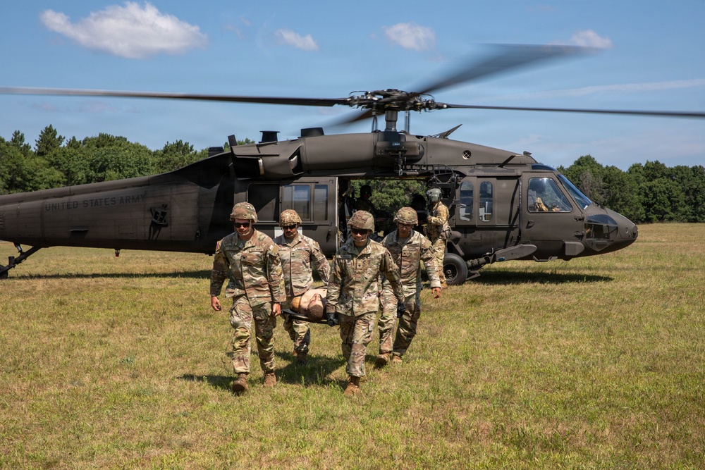 Four U.S. Army soldiers in camouflage uniforms carry a casualty on a litter (stretcher) away from a UH-60 Black Hawk helicopter on an open grassy field. The helicopter, marked 'UNITED STATES ARMY,' has its rotors spinning and doors open, suggesting an active medical evacuation (MEDEVAC) exercise or operation. Additional personnel are visible near the aircraft in the background.