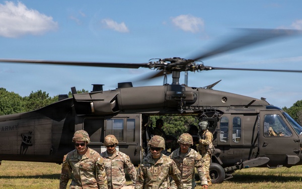 Four U.S. Army soldiers in camouflage uniforms carry a casualty on a litter (stretcher) away from a UH-60 Black Hawk helicopter on an open grassy field. The helicopter, marked 'UNITED STATES ARMY,' has its rotors spinning and doors open, suggesting an active medical evacuation (MEDEVAC) exercise or operation. Additional personnel are visible near the aircraft in the background.