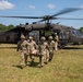 Four U.S. Army soldiers in camouflage uniforms carry a casualty on a litter (stretcher) away from a UH-60 Black Hawk helicopter on an open grassy field. The helicopter, marked 'UNITED STATES ARMY,' has its rotors spinning and doors open, suggesting an active medical evacuation (MEDEVAC) exercise or operation. Additional personnel are visible near the aircraft in the background.
