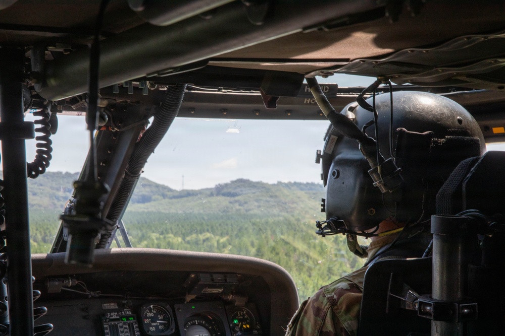 The image shows the interior of a military helicopter cockpit with a pilot or crew member visible in the right seat, wearing a flight helmet and camouflage uniform. The helicopter is in flight over a densely forested, hilly terrain under a partly cloudy sky. The cockpit instrumentation panel is partially visible in the lower foreground.