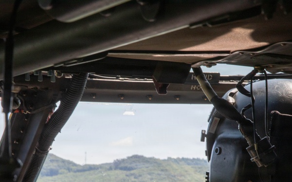The image shows the interior of a military helicopter cockpit with a pilot or crew member visible in the right seat, wearing a flight helmet and camouflage uniform. The helicopter is in flight over a densely forested, hilly terrain under a partly cloudy sky. The cockpit instrumentation panel is partially visible in the lower foreground.