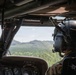The image shows the interior of a military helicopter cockpit with a pilot or crew member visible in the right seat, wearing a flight helmet and camouflage uniform. The helicopter is in flight over a densely forested, hilly terrain under a partly cloudy sky. The cockpit instrumentation panel is partially visible in the lower foreground.