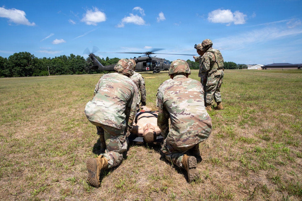 Military personnel in U.S. Army OCP (Operational Camouflage Pattern) uniforms are conducting a medical evacuation (MEDEVAC) training exercise on an open grassy field. Two soldiers are kneeling beside a simulated casualty lying on a litter/stretcher, while a third soldier appears to be giving instructions or directing the exercise. A UH-60 Black Hawk helicopter marked with a red cross (indicating medical/MEDEVAC designation) is landed in the background.