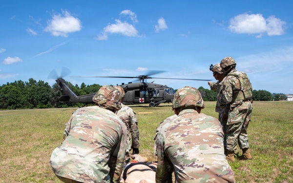 Military personnel in U.S. Army OCP (Operational Camouflage Pattern) uniforms are conducting a medical evacuation (MEDEVAC) training exercise on an open grassy field. Two soldiers are kneeling beside a simulated casualty lying on a litter/stretcher, while a third soldier appears to be giving instructions or directing the exercise. A UH-60 Black Hawk helicopter marked with a red cross (indicating medical/MEDEVAC designation) is landed in the background.