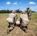 Military personnel in U.S. Army OCP (Operational Camouflage Pattern) uniforms are conducting a medical evacuation (MEDEVAC) training exercise on an open grassy field. Two soldiers are kneeling beside a simulated casualty lying on a litter/stretcher, while a third soldier appears to be giving instructions or directing the exercise. A UH-60 Black Hawk helicopter marked with a red cross (indicating medical/MEDEVAC designation) is landed in the background.