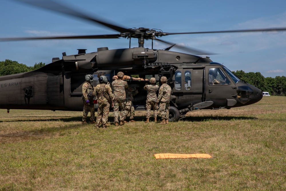 A group of U.S. Army soldiers are loading or unloading a casualty onto a UH-60 Black Hawk helicopter on a grassy field. The helicopter's rotors are spinning, indicating it is either landing or preparing for takeoff. This appears to be a medical evacuation (MEDEVAC) exercise or operation, evidenced by the red cross marking on the aircraft and the coordinated team effort.